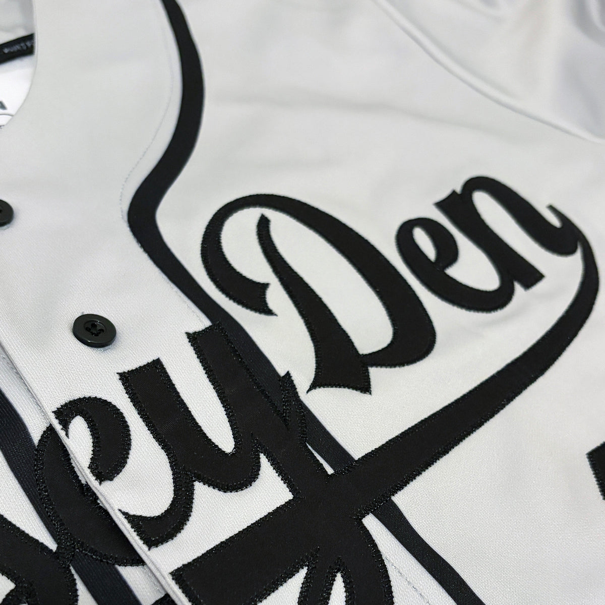 Close-up of a white baseball-style jacket with large black script lettering across the chest and black trim details.