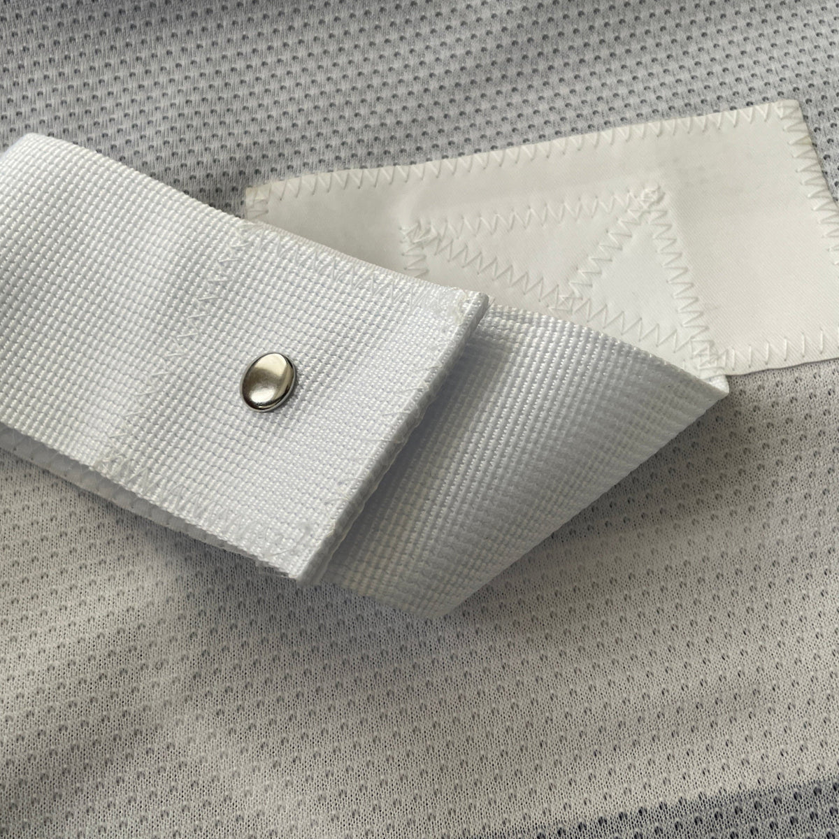 Close-up of a white adjustable fabric strap with a metal snap button and a white hook-and-loop fastening patch, laid on a gray mesh-textured surface.