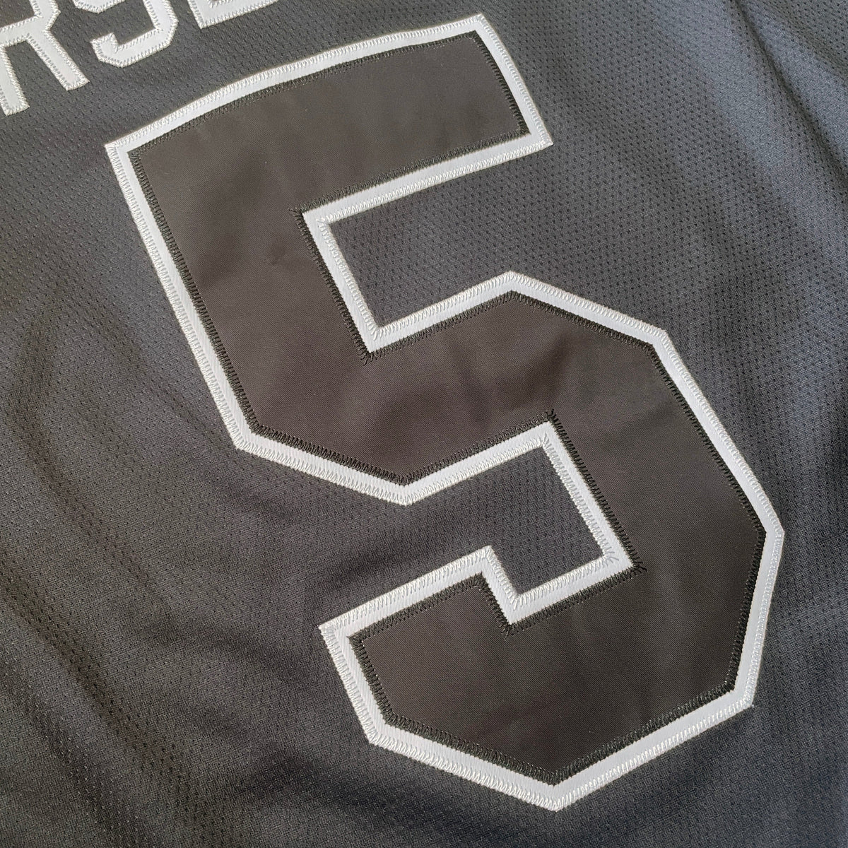 Close-up of the back of a dark gray athletic jersey with white lettering reading 'JERSEY DEN' and a large white-outlined number 5.