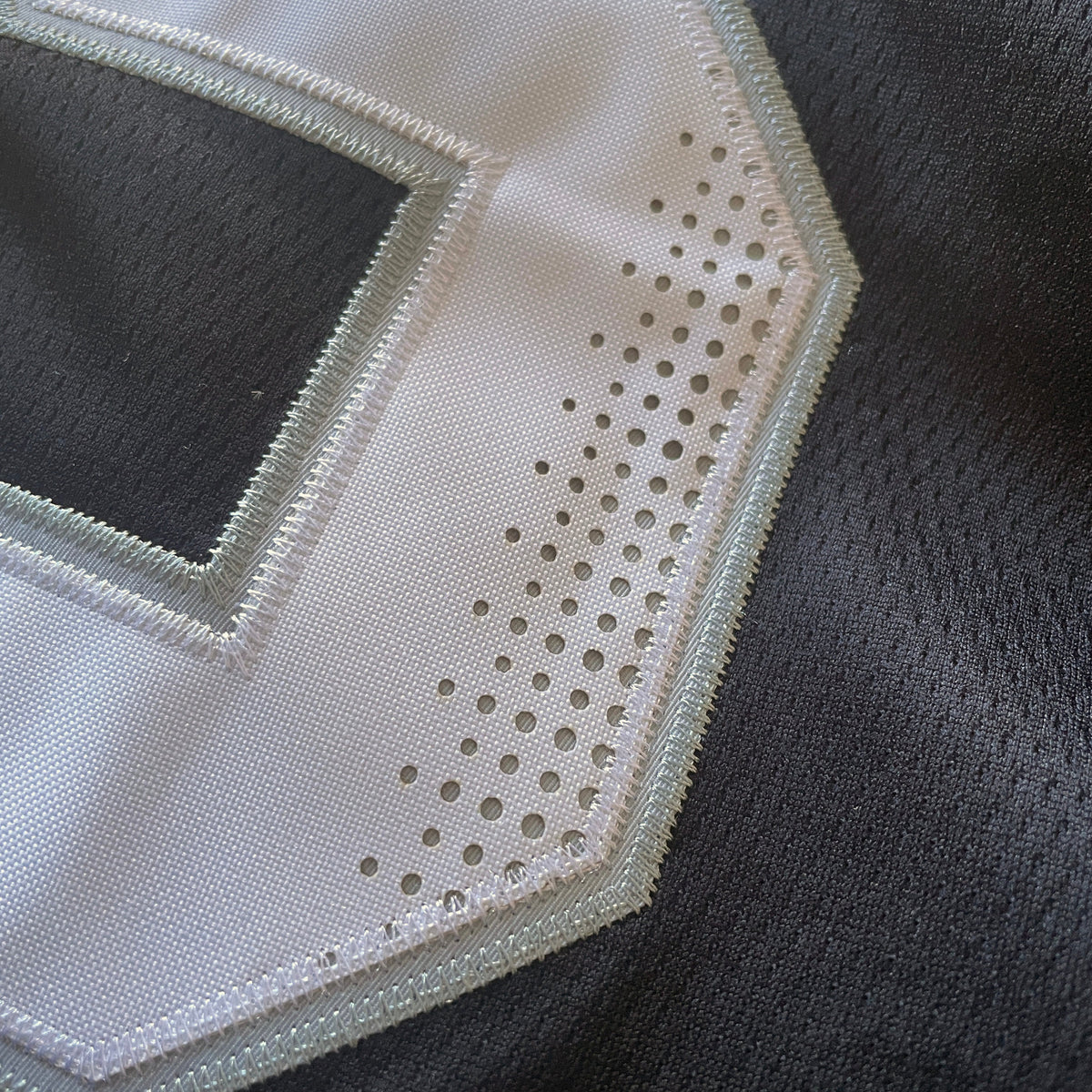 Close-up detail of a white embroidered letter patch with perforated circular cutouts and a stitched border, attached to a dark gray mesh fabric.