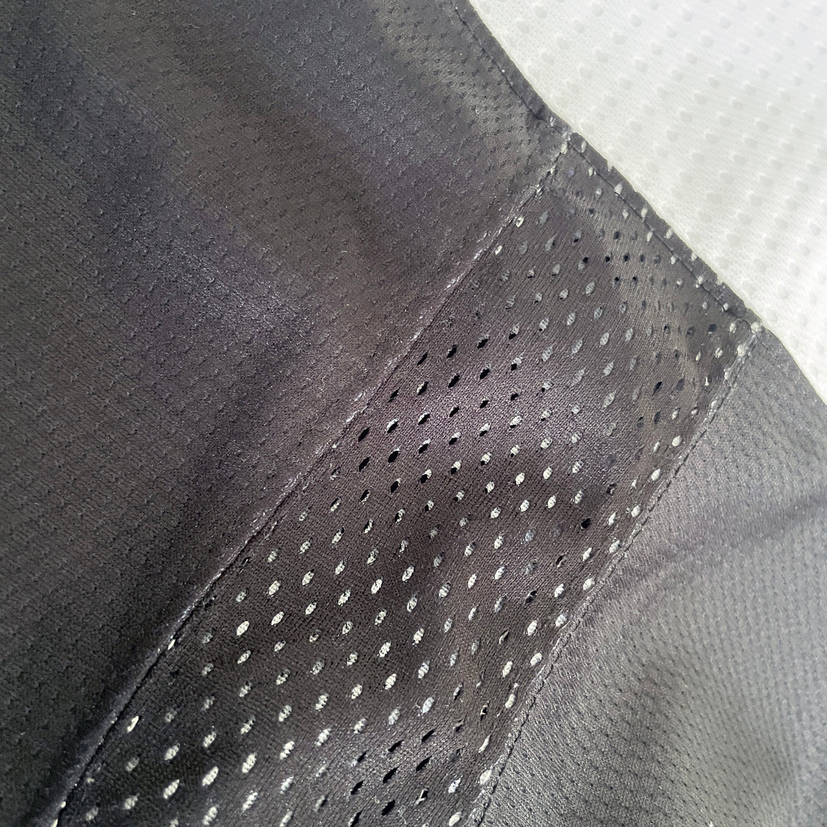 Close-up of a dark gray athletic mesh fabric with perforated holes, showing a stitched seam and texture against a white background.