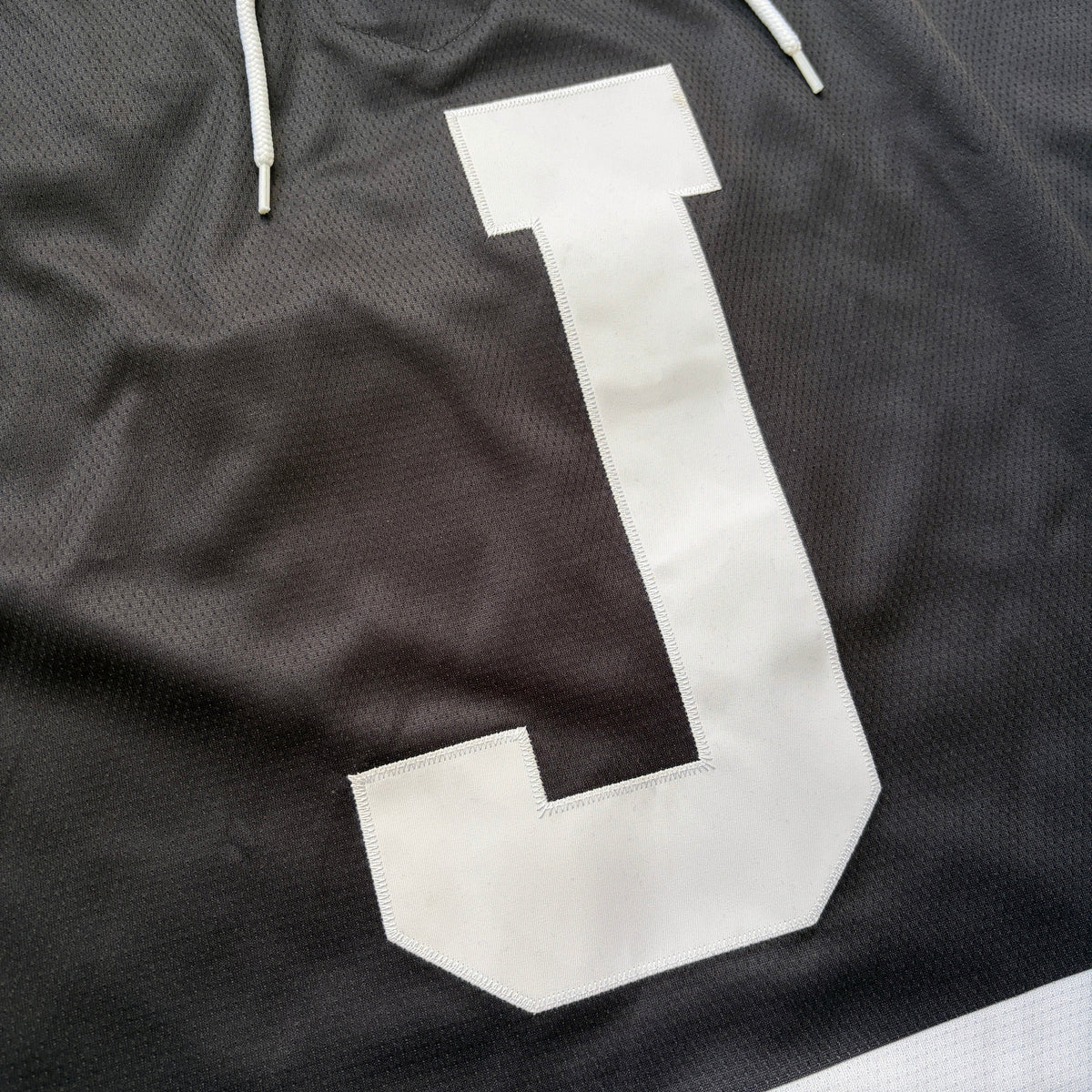 Close-up of a black mesh drawstring hoodie or sweatshirt with a large white letter J on the front and white drawstrings at the top.