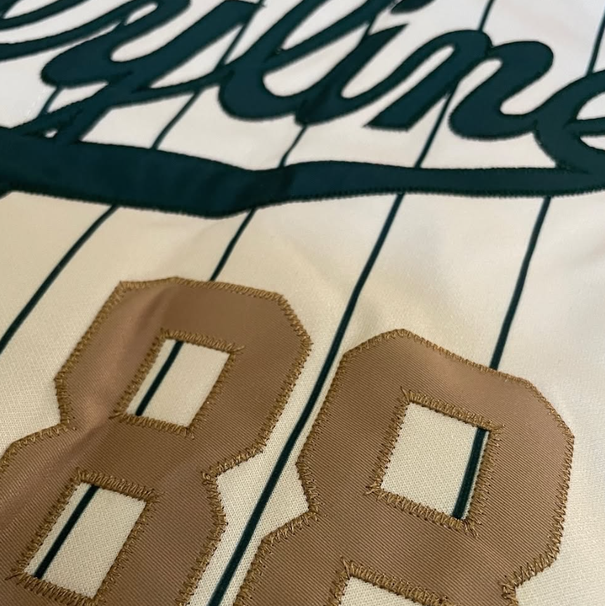 Close-up of a baseball-style jersey with dark teal lettering and pinstripes on an off-white fabric, featuring a large tan embroidered number 88.
