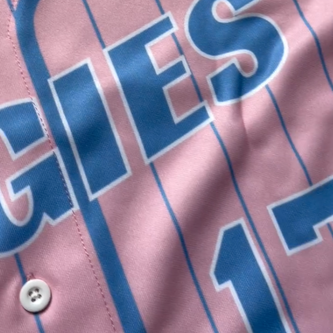 Close-up of a pink baseball jersey with blue pinstripes and large blue lettering outlined in white and a white button.